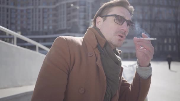 선글라스를 입고 도시 거리에 서 있는 갈색 코트에서 잘생긴 남자의 초상화. 남자 담배 흡연. 매력적인 남자의 여가. 배경의 도시 풍경. 슬로우 모션 — 비디오