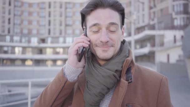 Retrato de un hombre guapo y exitoso de abrigo marrón parado en la calle de la ciudad hablando por teléfono móvil. Paisaje urbano en el fondo. Habitante de la ciudad moderna — Vídeo de stock