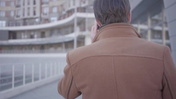 Vue arrière d'un bel homme en manteau marron marchant dans la rue de la ville parlant par téléphone portable. Paysage urbain en arrière-plan. Un citadin moderne. Caméra suit le gars — Video