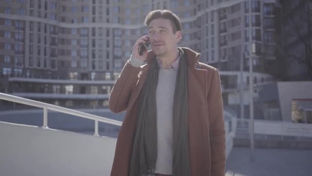 Porträt eines gutaussehenden, selbstbewussten Mannes in braunem Mantel, der auf der Straße steht und mit dem Handy telefoniert. Stadtbild im Hintergrund. moderner Großstadtbewohner. Kamera bewegt sich — Stockvideo