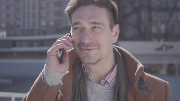 Retrato del exitoso hombre guapo y confiado en abrigo marrón parado en la calle de la ciudad hablando por teléfono celular. Paisaje urbano en el fondo. Habitante de la ciudad moderna. Cámara se mueve alrededor — Vídeos de Stock