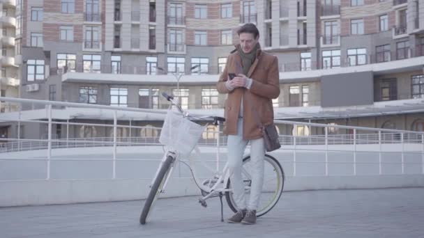 Портрет красивого мужчины, печатающего на клетке, стоя с велосипедом на фоне городской архитектуры. Парень отдыхает в хорошую солнечную погоду на открытом воздухе . — стоковое видео