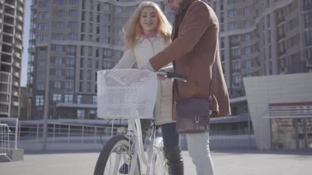 Красивый мужчина в коричневом пальто учит свою девушку кататься на велосипеде по городу, оба смеются. Отдых красивой пары горожан. Активный образ жизни — стоковое видео
