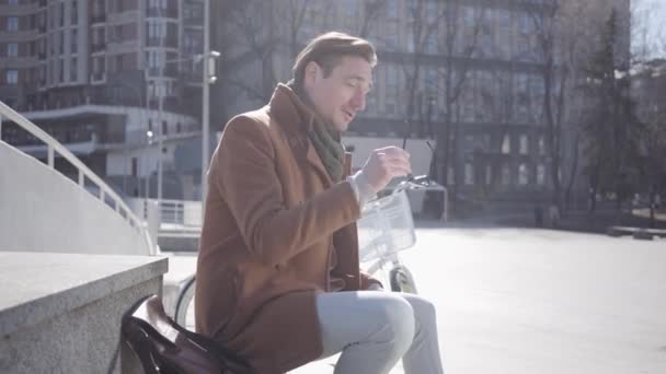 Портрет притягательного стильного мужчины в солнечных очках, сидящего на фоне городской архитектуры . — стоковое видео