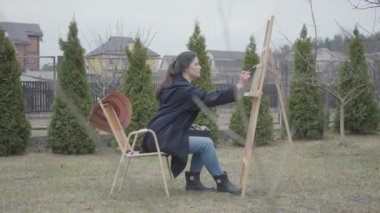 Güzel genç kız ressam bir resim çizim ahşap sehparın önünde oturuyor. Rahat kapanış kadın sanatçı zaman açık havada harcıyor. Yaratıcılık, kendi kendine ifade. Kamera doğru hareket ediyor. Yan görünüm