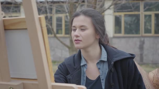 Porträtt framgångs rik vacker ung flicka målare sitter framför trä staffli tittar runt. Kvinnlig konstnär i casual stängning utomhus. Kreativitet, vila utanför, vackert landskap. — Stockvideo