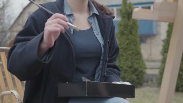 Una giovane donna dipinge un dipinto su una tela, in piedi dietro un cavalletto, in un laboratorio atmosferico nel cortile. Ispirazione. Talento. Accademia d'arte o scuola di disegno . — Video Stock