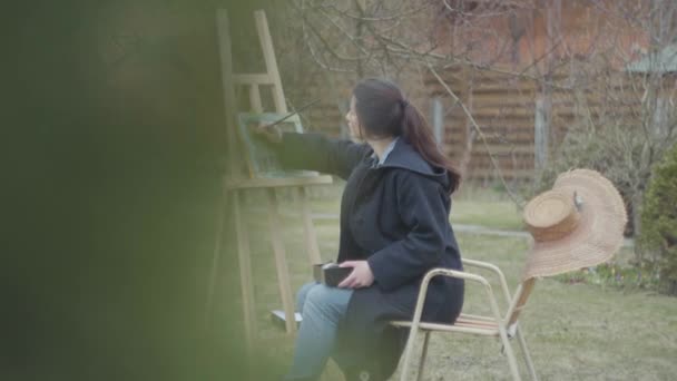 Pewna młoda dziewczyna malarza siedzi przed drewnianym sztalierem rozglądając się. Kobieta artystka w nieformalne zamknięcie siedzi na zewnątrz. Kreatywność, odpoczynek na zewnątrz, piękny krajobraz. Inspiracji. Talent. — Wideo stockowe