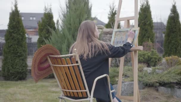 Widok z tyłu młodego malarza dziewczyna siedzi przed drewnianym sztalierem rysunek obraz i palenie. Kobieta artystka w swobodnym zamknięciu spędza czas na zewnątrz, obrazując krajobraz. Koncepcja kreatywności — Wideo stockowe