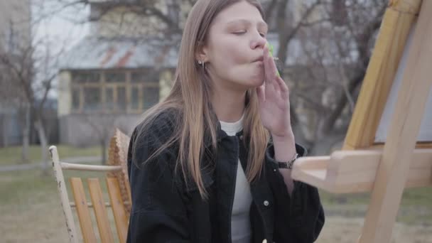 Portrét hezkě mladé kuřácké dívky na plátně, když sedí na dvoře venku. Úspěšný umělec vášnivou okupaci. Série skutečných lidí. — Stock video