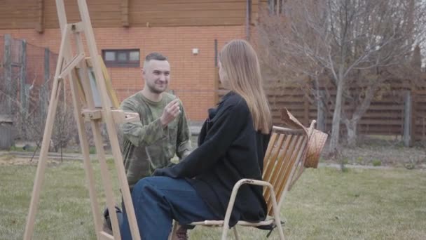 Ładna młoda kobieta malarza siedzi przed drewnianym sztalierem na zewnątrz. Szczęśliwy człowiek umieścić pierścień na palec Pani. Deklaracja miłości. — Wideo stockowe