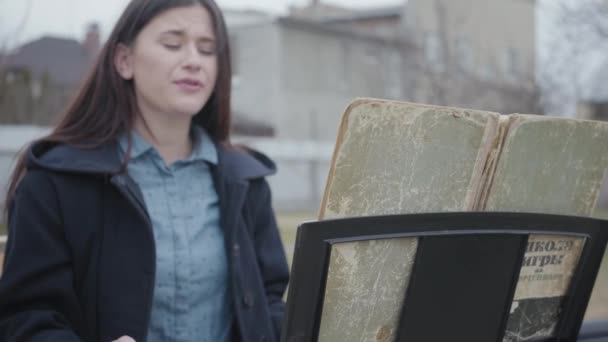 Portret udany piękna brunetka gra na syntezator i śpiewa siedząc na podwórku na zewnątrz. Młoda, udana kobieta jest chętna do zawodu. Seria prawdziwych ludzi. — Wideo stockowe