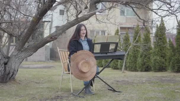 Επιτυχημένη ταλαντούχα όμορφη μελαχρινή κοπέλα που παίζει συνθεσάιζερ και τραγουδά ενώ κάθεται κάτω από το δέντρο στην αυλή του εξωτερικού χώρου. Ρομαντική, διαλογισμός, ηλεκτρονική μουσική — Αρχείο Βίντεο
