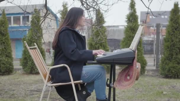 Vacker brunett tjej brinner för att spela synthesizer och sjunger medan du sitter under trädet i trädgården utomhus. Romantisk, meditation, elektronisk musik — Stockvideo