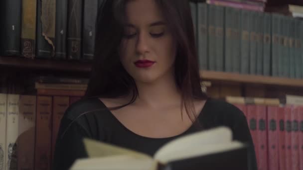 Retrato de un joven estudiante serio con maquillaje brillante leyendo un libro de pie frente a una estantería en una biblioteca. Ocio de mujer atractiva inteligente . — Vídeos de Stock