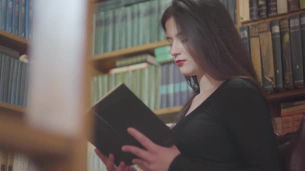 Retrato de chica morena leyendo un libro en la biblioteca sobre el fondo de las estanterías . — Vídeos de Stock