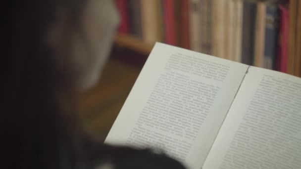 Vista posterior de la joven estudiante leyendo un libro en una biblioteca de cerca. La chica está pasando página del libro de pie delante de la estantería. Ocio de mujer atractiva inteligente — Vídeos de Stock