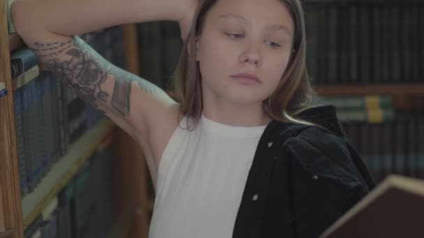 Retrato de una hermosa joven con un tatuaje en el libro de lectura del hombro en la biblioteca de cerca. Muchacha atractiva con el libro de pie delante de la estantería. Concepto de conocimiento, educación — Vídeos de Stock