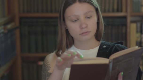 Πορτρέτο του χαριτωμένο κορίτσι με ένα τατουάζ στον ώμο διαβάζοντας βιβλίο στη βιβλιοθήκη κοντά. Ελκυστικό νεαρό κορίτσι με το βιβλίο να στέκεται μπροστά στη βιβλιοθήκη. Έννοια της γνώσης, εκπαίδευση — Αρχείο Βίντεο