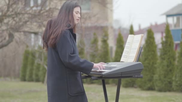 Portrait beautiful brunette girl passionate about playing synthesizer and sings while standing under the tree in the backyard outdoors. Romantic, meditation, electronic music — Stok video