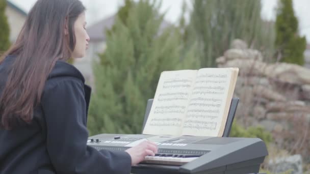 สาวผมสีน้ําตาลสวยหลงใหลเกี่ยวกับการเล่น synthesizer และร้องเพลงนั่งอยู่ใต้ต้นไม้ในสวนหลังบ้านกลางแจ้ง โรแมนติก การทําสมาธิ เพลงอิเล็กทรอนิกส์ — วีดีโอสต็อก