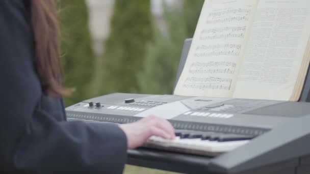 Chica no reconocida apasionada por jugar sintetizador y canta sentado bajo el árbol en el patio trasero al aire libre. Romántico, meditación, música electrónica — Vídeos de Stock