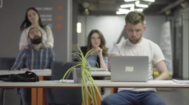 Beyaz tişört konsantre genç adam laptop ile çalışmalarını bitirmek ve ofiste yürüyor. Adam ve kadın boynları yoğurma, kız arka planda beardie Masaj yapma