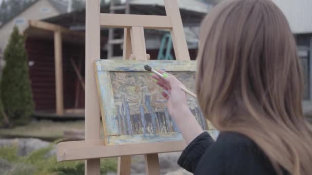 Профессиональная молодая девушка рисует на холсте, сидя на заднем дворе. Успешный художник, увлеченный своей профессией. Настоящие люди. Вид сзади . — стоковое видео