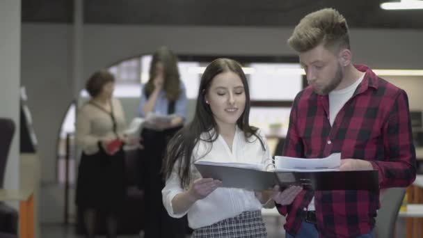 Young pretty woman and bearded man discussing project standing in the office with big paper folder. Their female colleagues in the background talking about documents in their hands. Working moments — Stock Video