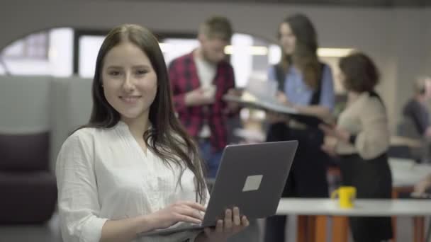 Retrato de la mujer atractiva y confiada mirando a la cámara sosteniendo netbook en las manos frente a figuras borrosas de colegas discutiendo sobre el proyecto juntos sosteniendo una carpeta grande — Vídeo de stock