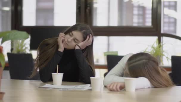 Attractive girl sitting at the table propped head with hand in the office, mixing her coffee, her friend leaning on the desk sitting near. Women are tired and bored. Camera moves left — Stock Video