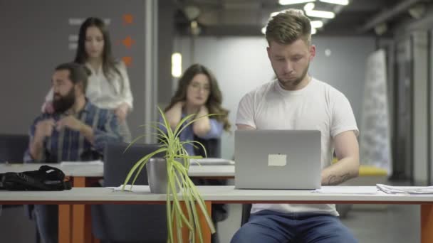 The concentrated young man in white t-shirt working with laptop sitting at the table in the modern office in the foreground. The man and woman kneading their necks, girl making massage to beardie — Stock Video