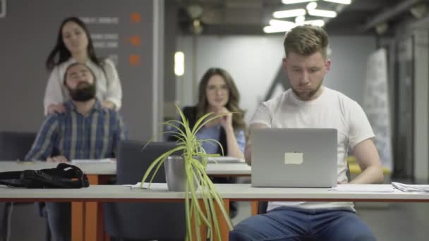 De geconcentreerde jonge man in wit t-shirt afwerking zijn werk met de laptop en loopt weg in het kantoor. De man en vrouw kneden hun nek, meisje het maken van massage te beardie op de achtergrond — Stockvideo