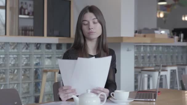 Attraktive junge Frau im schwarzen Kleid, die am Tisch in einem modernen Café sitzt und Dokumente liest. weiße Teekanne und Tasse stehen auf dem Tisch. Fernarbeit. Freiberufler. Serie "echte Menschen". — Stockvideo