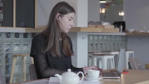 Attractive young woman in black dress sitting at the table in modern cafe working on her netbook. White teapot and cup are at the table. Lady is working in cafe. Remote work. Freelancer. — Stock Video
