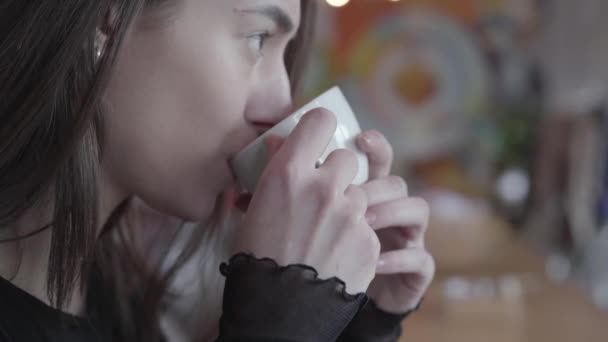 Primer plano retrato de una joven sentada a la mesa en la cafetería moderna bebiendo su té de taza blanca. A la bella dama le gusta su bebida. Serie de personas reales . — Vídeo de stock