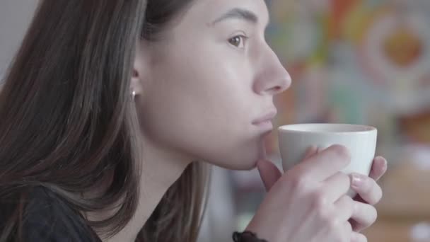 Zavřete portrét přemýšlivější mladé Brunetky, která pije čaj nebo kávu z bílého poháru, který odpočívá v kavárně nebo restauraci. Ta holka má ráda víkend nebo odpočinek. Série skutečných lidí. — Stock video