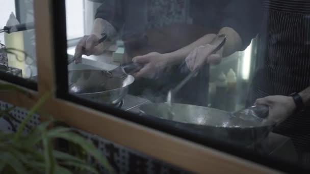 Las manos de los cocineros preparando la comida detrás del vidrio en el restaurante moderno de cerca. Dos hombres friendo comida en sartenes. Movimiento lento — Vídeo de stock