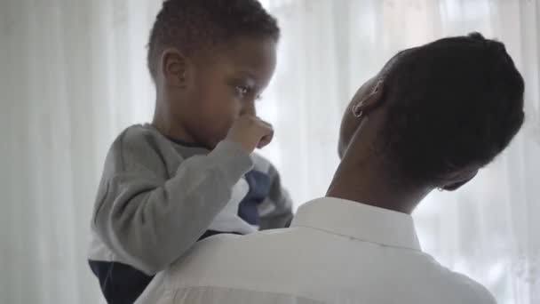 Mujer de estilo afroamericano en blusa blanca tomando en las manos su divertido niño juguetón en acogedora sala de estar. Relación madre e hijo. Una familia feliz . — Vídeos de Stock