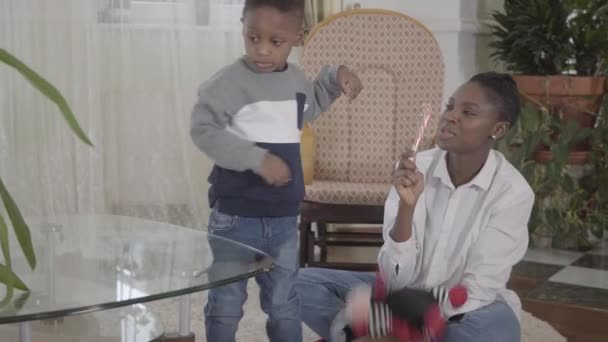 Σίγουρη όμορφη αφρικανική Αμερικανίδα που παίζει με το μικρό αστείο γιο της στο φιλόξενο σαλόνι. Σχέση μαμά και γιος. Μια ευτυχισμένη οικογένεια. — Αρχείο Βίντεο