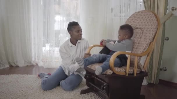 Alegre linda mujer afroamericana jugando con su pequeño hijo lindo y divertido sentado en una mecedora en la sala de estar. Relación madre e hijo. Una familia feliz . — Vídeo de stock