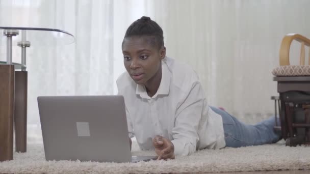 Πορτραίτο ελκυστική Afican αμερικανική γυναίκα επικεντρώθηκε δουλεύοντας μαζί της μέχρι την ημέρα laptop βρίσκεται σε πατάκια στο πάτωμα στο δωμάτιο στο σπίτι. Ελεύθερος επαγγελματίας. Απομακρυσμένη εργασία. — Αρχείο Βίντεο