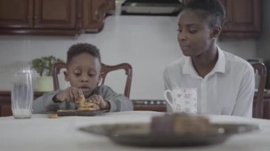 Çekici Afrikalı Amerikalı kadın masanın tarafından küçük oğlu ile oturma plaka üzerinde kurabiye oynuyor. İlişki anne ve oğul. Mutlu bir aile. Aile sağlıklı kahvaltı.