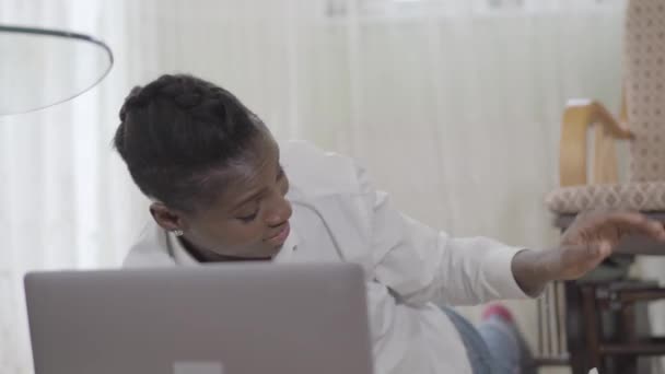 Афроамериканець жінка працює на ноутбуці лежачи на килимі і її маленький милий син кинув іграшку на неї — стокове відео