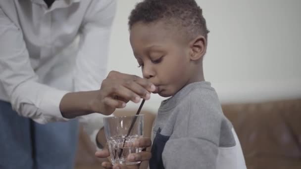 Zorgzame moeder helpen weinig Afro-Amerikaanse jongen om water te drinken uit glas met buis. Relatie moeder en zoon. Een gelukkige familie. — Stockvideo