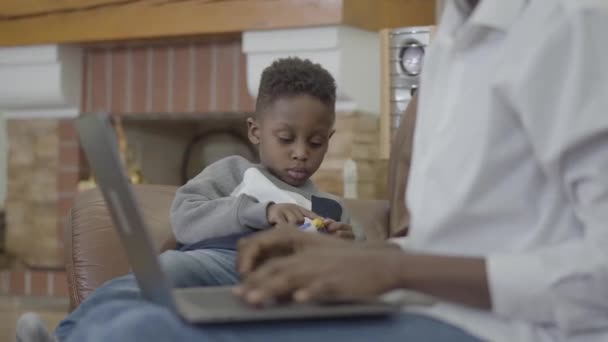 노트북에 작업 귀여운 아프리카계 미국인 여자와 아늑한 거실에서 장난감을가지고 노는 그녀의 작은 귀여운 아들. 아들과 관계 엄마. 행복 한 가족. — 비디오