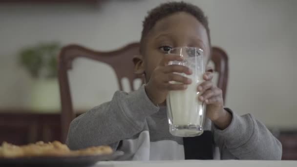 居心地の良いリビングルームの背景にガラスからテーブルに座って、ミルクを飲むかわいいアフリカ系アメリカ人の少年の肖像 — ストック動画