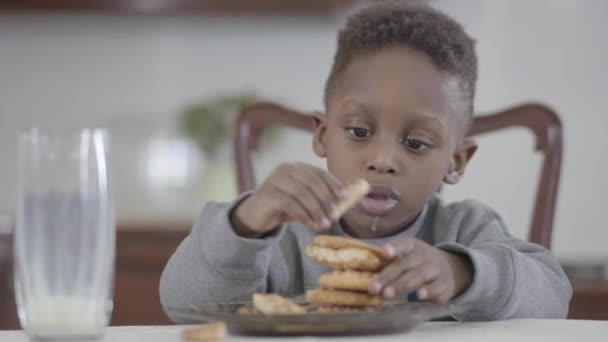 Портрет симпатичного африканского мальчика-американца с грязными губами после того, как молоко делает пирамиду печенья сидя за столом в столовой — стоковое видео