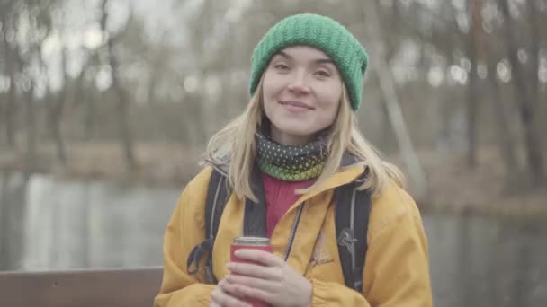 Portrait of confident cute young woman in green hat and yellow coat drinking tea or coffee from the thermos in the park looking in camera close up. Positive lady rests outdoors. — Stock Video
