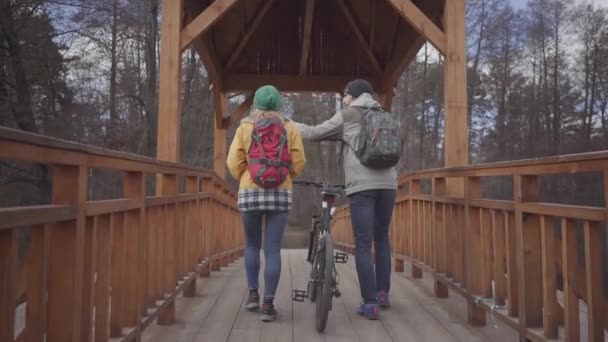 Twee jonge toeristen lopen op de brug in het park met rugzakken op hun rug. De man leidt de fiets en de vrouw in fel geel jasje wandelingen in de buurt. Happy Lovers rusten buitenshuis. — Stockvideo